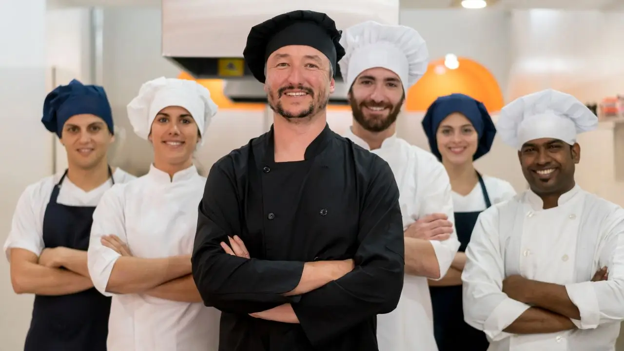 Team of chefs in a kitchen, featuring a head chef in a black uniform and five assistants in white and blue chef hats, representing Culinary Collective ATL's celebrity chef services.