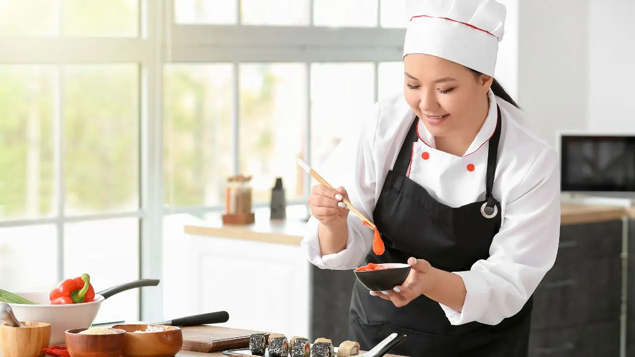 Chef in white uniform and hat carefully plating a dish with artistic garnishing in a bright kitchen, showcasing culinary creativity and attention to detail.