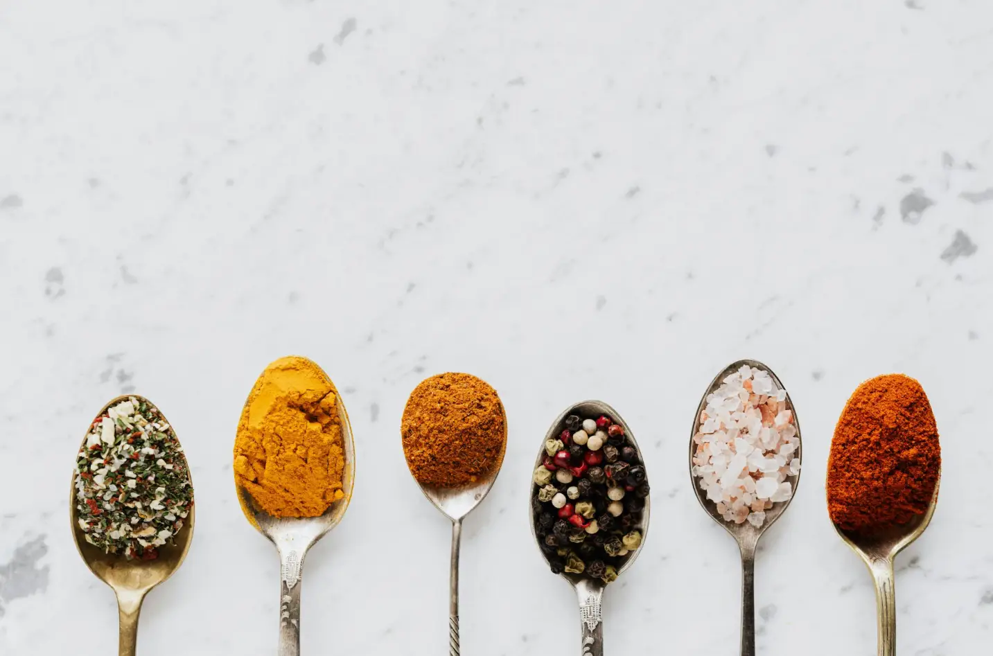 Assorted spices in silver spoons, including herbs, turmeric, curry powder, black pepper, Himalayan salt, and paprika, set against a marble background, highlighting culinary diversity for private chef services.